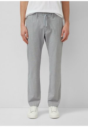 Mann trägt hellgraue Hose mit Kordelzug und weiße Turnschuhe, steht vor einem schlichten hellgrauen Hintergrund.