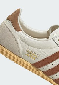 Biały skórzany sneaker z brązowymi zamszowymi akcentami. Posiada złote logo "adidas JAPAN", perforowane detale oraz teksturowaną gumową podeszwę.