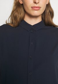 Tmavomodrá blúzka s gombíkovým zapínaním, špicatým golierom a hladkým, ľahkým materiálom. Má šesť gombíkov a uvoľnený strih.