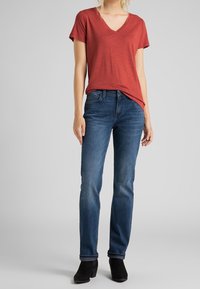Femme portant un T-shirt rouge en V, un jean bleu et des bottines noires se tenant contre un fond neutre.