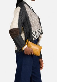 Chunky gebreide cardigan in grijs, crème en bruine texturen, gecombineerd met een blauwe wijde spijkerbroek. Houdt een kleine, felgele leren tas vast.