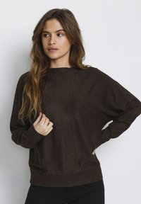 Maglione lungo marrone a maniche lunghe con una vestibilità ampia, polsini a coste e colletto a girocollo. Il tessuto ha una texture morbida con leggere variazioni.