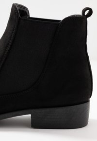 Černé semišové kotníkové boty s elastickými bočními panely, texturovaným povrchem, kulatou špičkou a nízkým, vrstveným černým podpatkem. Obsahují poutko.