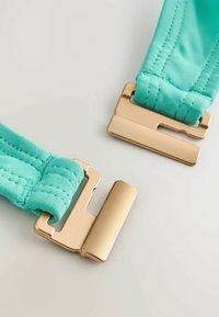 Arany fémkapocs türkizkék szövetpántokkal, világos felületen oldott állapotban bemutatva.