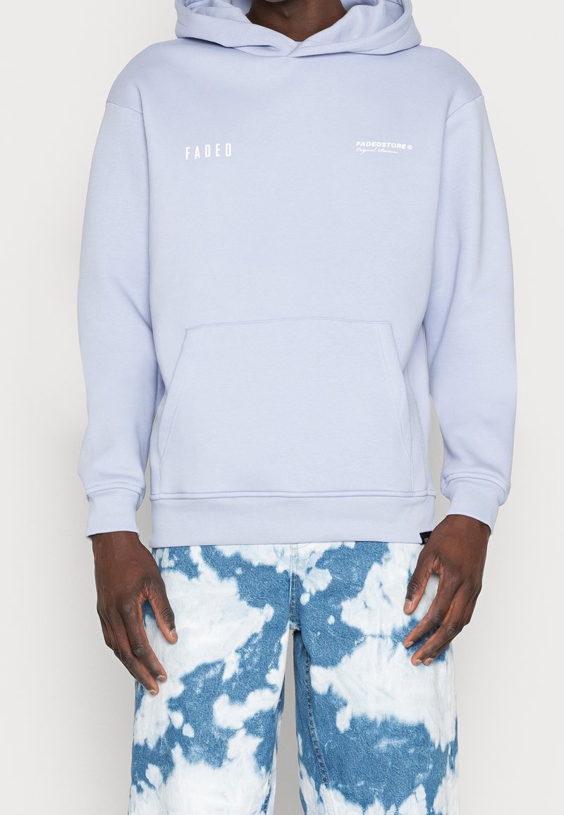 Felpa con cappuccio di colore viola chiaro con tasca frontale, decorata con testo bianco sul petto; abbinata a jeans tie-dye blu.