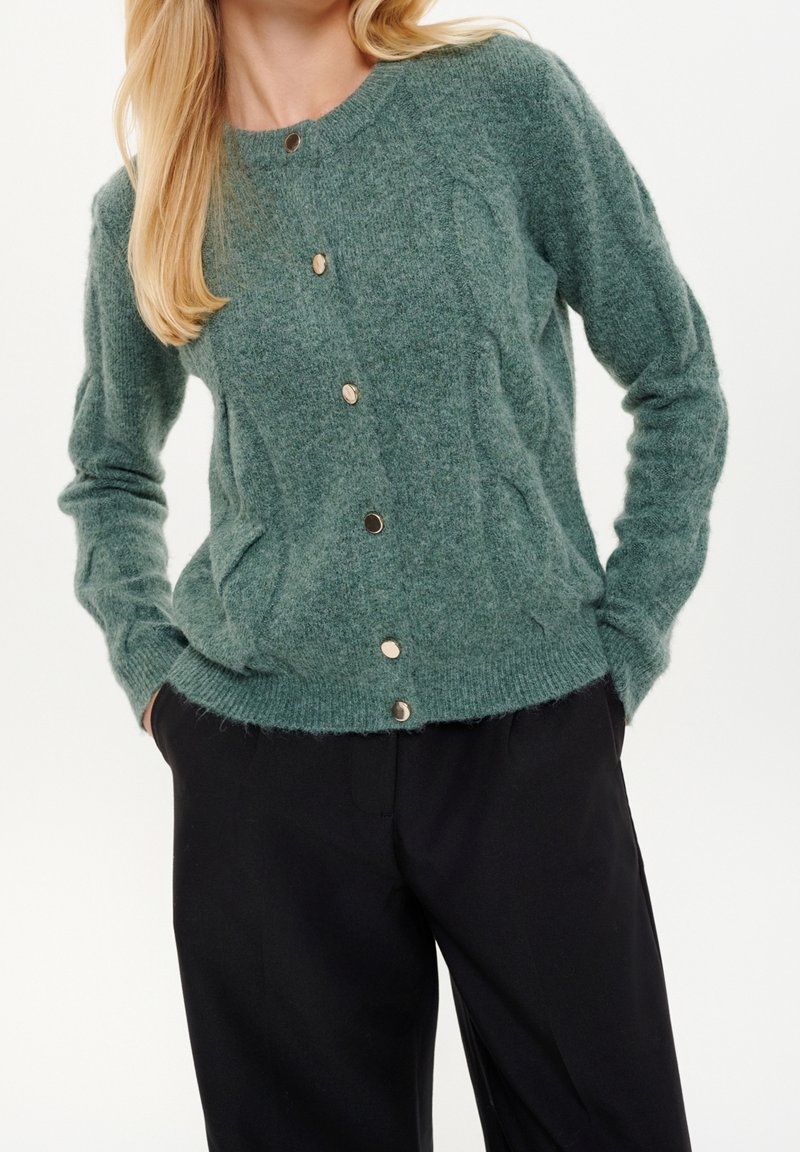 Femme portant un cardigan vert boutonné à manches longues et un pantalon noir, les mains dans les poches, sur un fond uni.