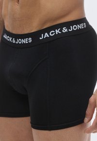 Melnas kokvilnas bokseršorti ar elastīgu jostu, kurā baltā krāsā redzams zīmola nosaukums "JACK & JONES". Gluda tekstūra ar redzamām šuvēm.