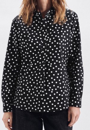 Kvinde iført en sort langærmet skjorte med hvide polka prikker, kombineret med mørke bukser, stående mod en ensfarvet baggrund.