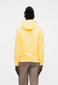 Κίτρινο φούτερ με κουκούλα και απαλή υφή, με τσέπη καγκουρό, συνδυασμένο με ανοιχτό καφέ παντελόνι ραμμένο. Πλάγια όψη δείχνει την κο υκούλα.