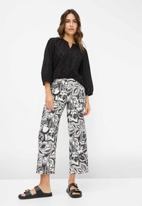 Zwarte blouse met knoopjes en een gemêleerd textuur, gecombineerd met wijde, zwart-witte bloemenbroek. Zwarte slides met gouden accenten.