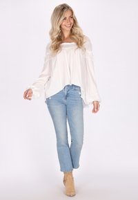Hvid, let bluse med blonde detaljer og rynkede ærmer, parret med lyseblå jeans og beige ankelstøvler. Casual look.