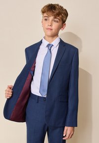 Niño con traje azul a cuadros, corbata azul claro y camisa blanca, sosteniendo abierta una parte de su chaqueta sobre un fondo beige liso.
