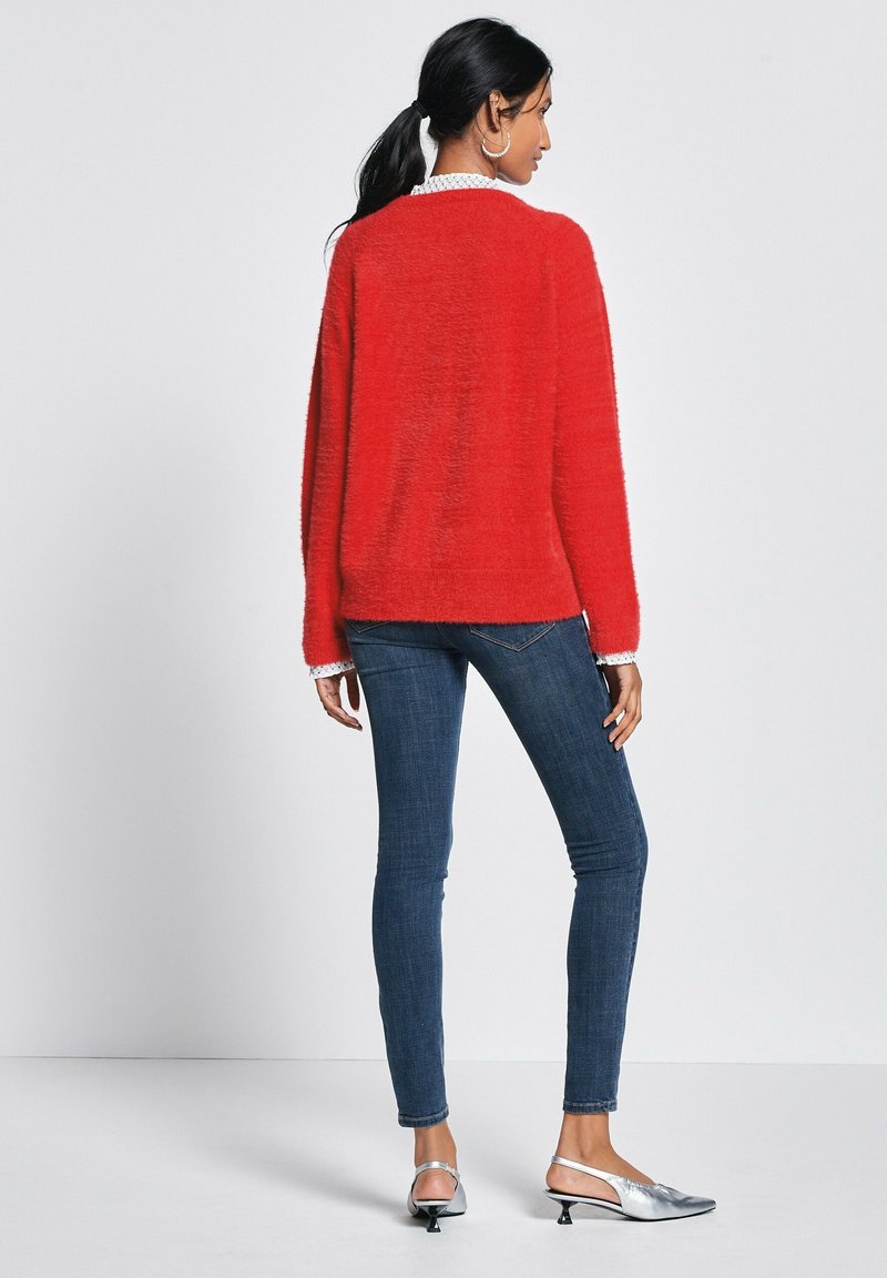 Czerwony, puszysty sweter o luźnym kroju, w połączeniu z ciemnoniebieskimi obcisłymi dżinsami i srebrnymi butami na odkrytej pięcie. Teksturowany materiał i swobodny fason.