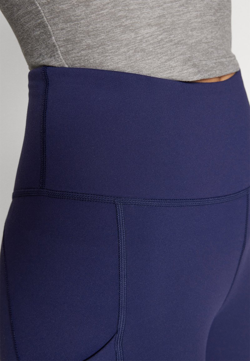 Hochgeschnittene marineblaue Leggings mit verstärkten Nähten, glatter Textur und einem breiten Bund, die zu einem hellgrauen Oberteil passen.