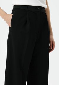 Zwarte op maat gemaakte broek met zijzakken, een enkele knoopsluiting, plooien en een gladde, gestructureerde stof.