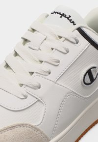 Άσπρο αθλητικό παπούτσι με υφασμένα κορδόνια, διάτρητες πλευρικές επιφάνειες, λογότυπο Champion στο πλάι και λεπτομέρεια από σουέντ στο μπροστινό μέρος.