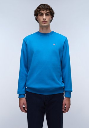Blauwe katoenen sweatshirt met ribgebreide manchetten en zoom, met een klein rood en wit logo op de borst. Model draagt marineblauwe broeken.