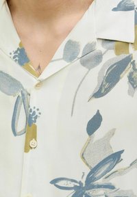 Chemise à manches courtes en tissu crème clair avec des motifs floraux verts et bleus, dotée d'un col pointu et de boutons beiges.