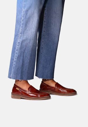 Personne portant un jean bleu droit et des mocassins en cuir marron brillant, debout sur un fond clair uni.