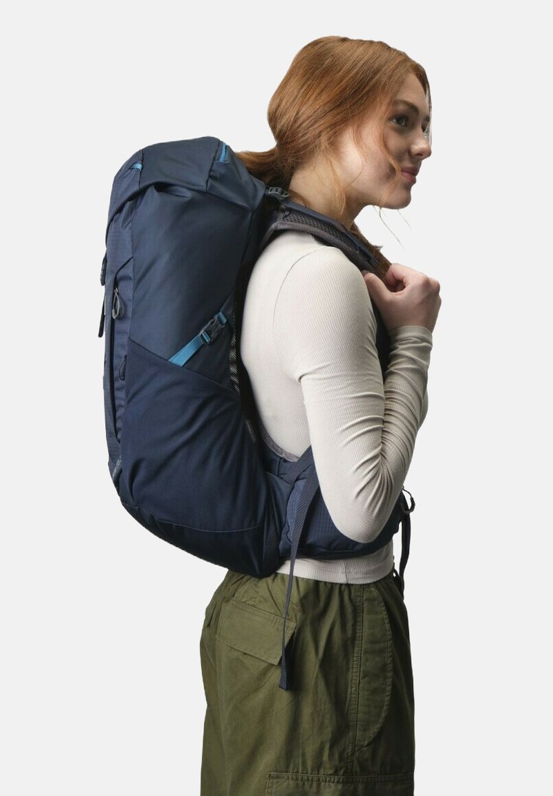 Sac à dos en tissu bleu marine avec bretelles rembourrées, poches latérales et boucles réglables, porté sur le dos d'une personne sur un fond blanc.