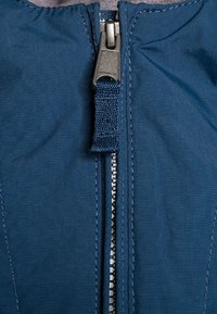 Chaqueta azul con cremallera que presenta una cremallera metálica y una lengüeta de tejido. La textura de la tela parece suave con costuras visibles a lo largo de las costuras.