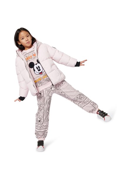 Bambino che indossa un puffer jacket rosa chiaro, una felpa di Topolino, pantaloni a fantasia e scarpe da ginnastica bianche, in equilibrio su una gamba contro uno sfondo bianco.