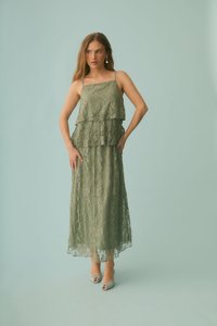 Femme debout devant un fond bleu clair, portant une longue robe en dentelle vert olive avec de fines bretelles et des volants superposés, posant avec une main sur la hanche.