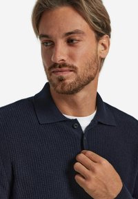 Mörkblå texturerad polo-shirt med krage, knappslå och ribbad detalj. Bärs över en vit T-shirt.