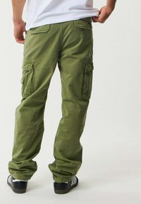Pantaloni cargo verde oliva con multiple tasche con pattina, vestibilità rilassata e una leggera texture stropicciata. Indossati con sneakers bianche e nere.
