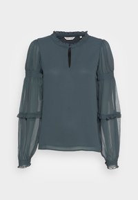 Blouse teal à manches longues avec un col à volants et des manches bouffantes transparentes. Possède un bouton à la devant dans une découpe en forme de clé et une texture subtile.