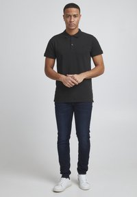 Camisa polo negra de manga corta, con tapeta de dos botones y un pequeño logo. Combinada con unos vaqueros azul oscuro y zapatillas blancas. Textura de tela suave.