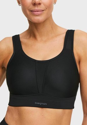 Vrouw draagt een zwarte sportbeha met brede bandjes en een glad, ondersteunend ontwerp met het merk "swegmark" op de voorste band.