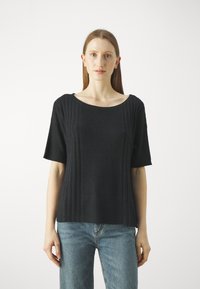 Čierny rebrovaný pletený t-shirt s krátkym rukávom a širokým výstrihom, v kombinácii s svetlo modrými džínsmi. Hladká textúra a voľný strih.