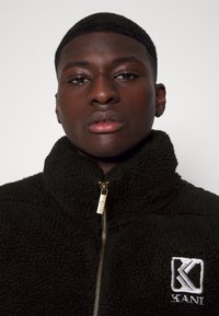 Jachetă din fleece neagră cu guler înalt, fermoar aurie și logo brodat pe pieptul stâng. Textura moale cu un model ușor reliefat.