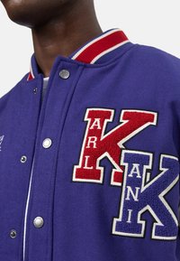 Lila ullvarsityjacka med röda och blå broderade bokstäver "K" och "ARKANI." Har tryckknappar och randig ribbad krage.