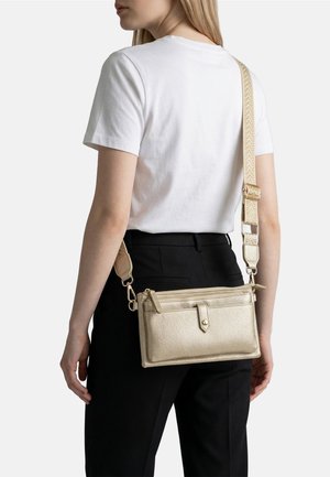 Donna che indossa una maglietta bianca e pantaloni neri con una borsa a tracolla dorata effetto texture, dotata di più scomparti con cerniera.