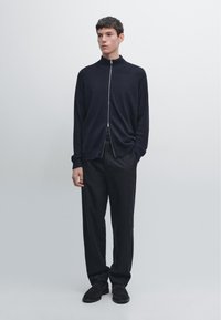 Granatowy kardigan na zamek wykonany z lekkiego dzianiny, zestawiony z ciemnymi, dopasowanymi spodniami. Charakterystyczny wysoki kołnierz i ściągacze na mankietach.