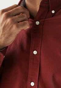 Camisa de botones color granate con cuello, con botones blancos y tejido texturizado. Primer plano mostrando una mano ajustando el cuello.