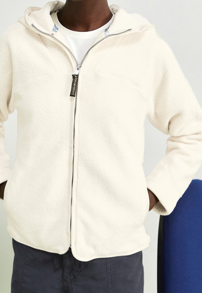 Persona che indossa una giacca in pile crema con zip, con le mani nelle tasche e una maglietta bianca sotto, in piedi accanto a una sedia blu.
