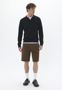 Camisa negra de manga larga con cuello en V, pantalones cortos marrones texturizados, calcetines blancos hasta el tobillo y mocasines negros, creando un atuendo en capas.