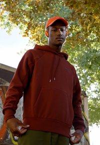 Burgundy huvtröja med en framficka och dragsko på huvan, ihop med en orange keps. Tyget verkar mjukt med en avslappnad passform.