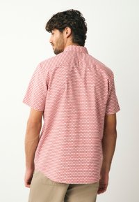 Chemise à manches courtes rouge avec un motif géométrique. Le tissu semble léger et texturé, avec un col classique et un ourlet arrondi.