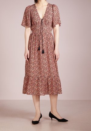 Mujer con vestido midi floral con mangas acampanadas, escote en V, lazo con borlas y zapatos de tacón bajo negros sobre fondo rosa pálido.