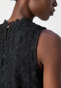 Gros plan sur l'épaule et le cou d'une personne portant un haut noir en dentelle sans manches avec des motifs floraux et une fermeture à boutons dans le dos.