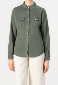 Personne portant une chemise boutonnée olive à manches longues avec deux poches poitrine et un pantalon écru sur un fond uni.