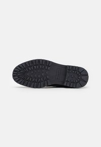 Suela de zapato de goma negra con surcos profundos para agarre, superficie texturizada y un detalle central con logo. Diseñada para tracción y durabilidad.