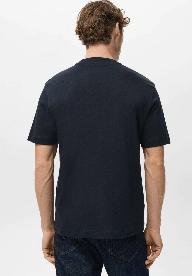 Uomo con capelli ricci che indossa una semplice maglietta a maniche corte blu navy scuro e jeans scuri, di spalle contro uno sfondo chiaro.