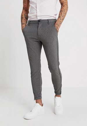 Mann trägt schmal geschnittene graue Hose mit Seitenstreifen, weiße Sneakers und ein weißes kurzärmeliges Hemd, tätowierte Arme sichtbar, Hände in den Taschen.