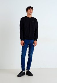 Sweat-shirt noir avec des poignets côtelés et un petit logo, associé à un jean bleu et des baskets noires. Le mannequin se tient devant un fond uni.