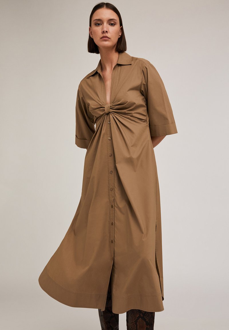 Abito camicia in cotone marrone con maniche corte, dettaglio anteriore annodato e chiusura con bottoni; lunghezza al ginocchio con una silhouette rilassata.